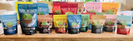 A row of Flavor Seed 5 oz resealable seasoning pouches in blue, green, red, yellow and teal, standing side by side on a wooden kitchen counter.