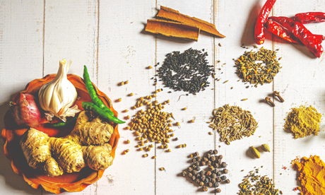 Whole spices laid out on white painted wood: cinnamon sticks, dried red chillies, cumin, coriander, fennel and mustard seeds, cloves, cardamom and black peppercorns, beside a bowl of garlic, red onion, green chillies and fresh ginger.