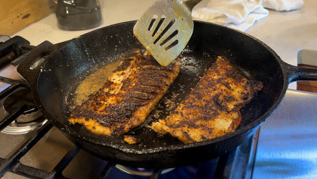 Two white fish fillets with a dark spice crust cooking in butter in a black cast iron skillet on a stovetop, a slotted spatula lifting the edge of one