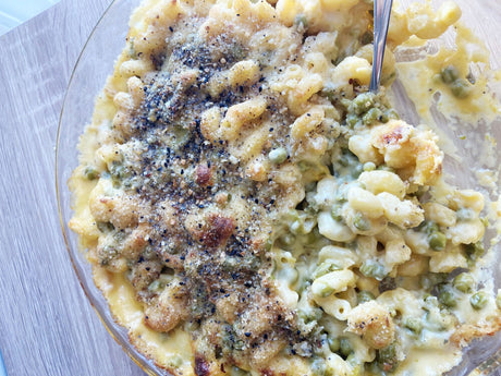 A glass baking dish of baked macaroni and cheese studded with green peas under a browned breadcrumb and coarse black pepper crust, with a spoon dug into the right side, on a pale wood board.
