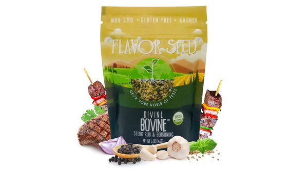 Flavor Seed Divine Bovine organic steak rub and seasoning pouch on a white background, with a grilled steak, two beef and vegetable kebabs, half a red onion, garlic, basil and black peppercorns in a wooden spoon around it.