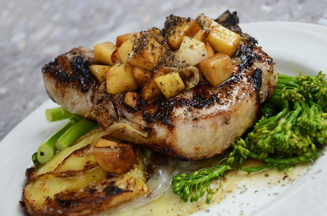 A thick seared bone-in pork chop on a white plate topped with browned diced apple and onion in a pan sauce, with broccolini and a roasted potato alongside