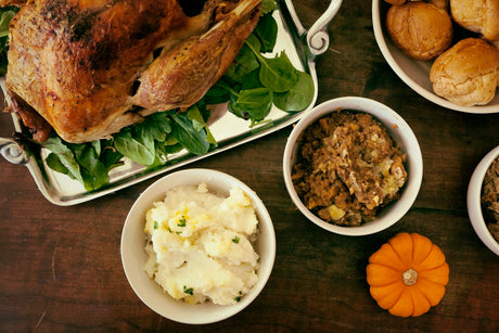 An overhead view of a Thanksgiving table: a whole roasted turkey on a silver platter over fresh greens, a bowl of mashed potatoes with melting butter, a bowl of stuffing, dinner rolls and a small orange pumpkin on a dark wood table.