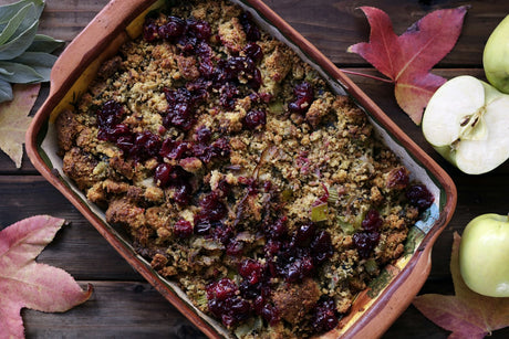 A rectangular terracotta baking dish of baked cornbread dressing, its golden crumb flecked with green celery and scattered with dark red cranberries, on a dark wood table with autumn leaves and green apples beside it.