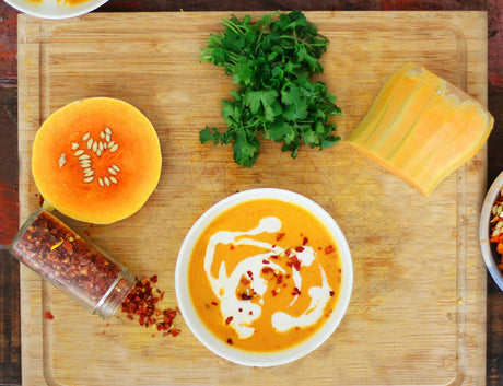 Overhead view of a white bowl of deep orange butternut squash soup with a cream swirl and crushed red pepper, on a wood board beside a halved butternut squash, fresh cilantro and a jar of red pepper flakes