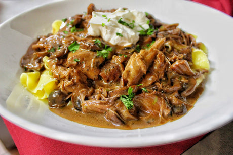 A white bowl of beef stroganoff: shredded beef and sliced mushrooms in a creamy brown sauce over wide egg noodles, topped with sour cream and chopped parsley.