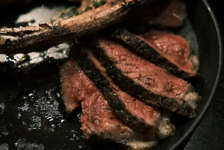 Slices of bone-in steak with a dark herb-and-pepper crust and a pink center resting in a cast iron skillet, the rib bone alongside