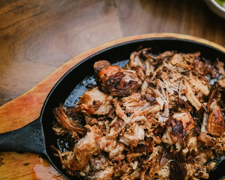 A cast iron skillet heaped with shredded pulled pork with crisp browned bark, on a warm wooden table.