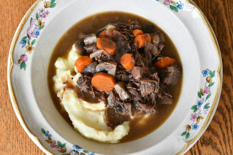 An overhead view of a floral-rimmed ceramic bowl holding mashed potatoes topped with shredded braised beef, sliced mushrooms, rounds of carrot and brown gravy, on a wooden table.