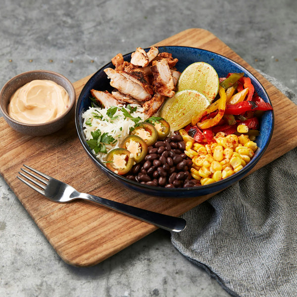 A blue bowl on a wooden board holding sliced seasoned chicken, white rice with cilantro, black beans, chilli-dusted corn, sliced pickled jalapenos, roasted red and yellow peppers and two lime halves, with a small bowl of pale orange sauce alongside