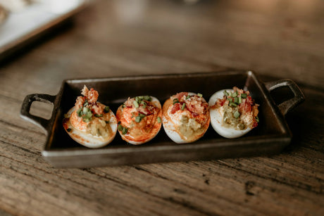 Four deviled eggs topped with crumbled bacon, chopped green onion and a dusting of paprika, in a dark rectangular serving dish on a weathered wooden table.