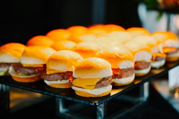 Rows of small cheeseburger sliders on a dark glass serving board, each a soft golden bun with a beef patty and a square of melted orange cheese, a few with tomato and lettuce showing.