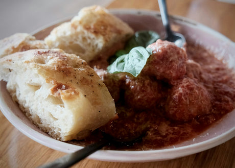 Meatballs in tomato sauce with a dusting of grated cheese and a fresh basil leaf in a shallow bowl, with three pieces of focaccia, a fork and a spoon, on a wooden table