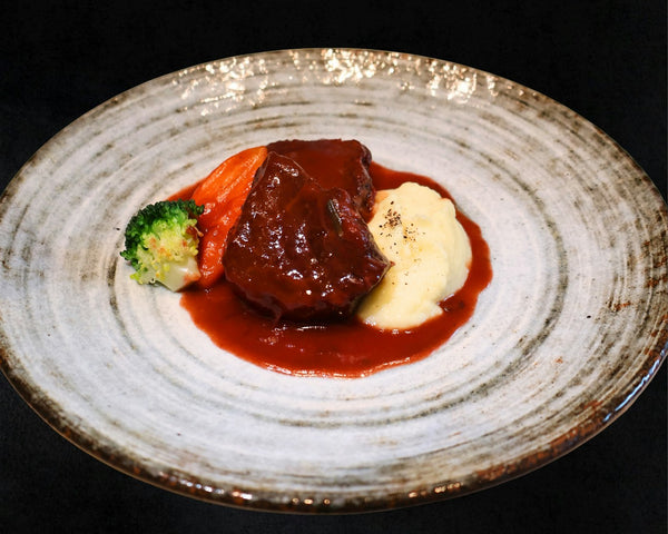 A braised beef chuck in glossy dark gravy on a speckled stoneware plate with mashed potato, a carrot and a broccoli floret